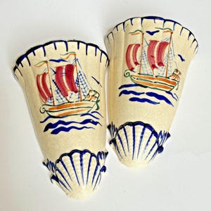 May include: Two cream-colored ceramic wall pockets with a crackle glaze finish. Each piece features a hand-painted sailing ship with red sails on blue waves. The top and bottom edges are decorated with a scalloped shell design in dark blue.