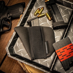 May include: A black holster with a textured weave, a black handgun, and a black magazine are displayed on a diamond-plate surface. Several gold-colored bullets and a carbon fiber wallet with a red band are also visible.
