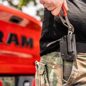 May include: A person wearing camouflage pants and a black shirt is holding a red and black pliers. The pliers are being inserted into a black carbon fiber holster attached to a belt.