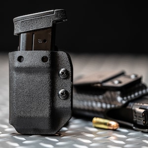 Może przedstawiać: Czarny uchwyt na magazynek z włożonym magazynkiem, zabezpieczony dwiema śrubami. Kabura na broń i nabój widoczne w tle. Uchwyt ma teksturowaną powierzchnię.
