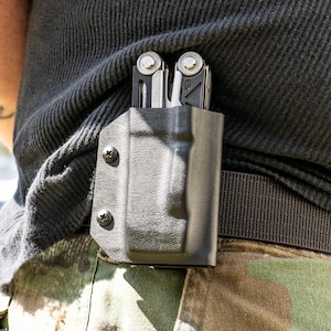 May include: A black multi-tool with a silver blade is secured in a black leather holster. The holster is attached to a black belt.