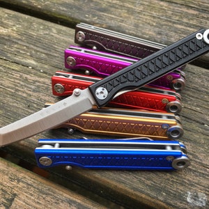 Könnte beinhalten: Eine Sammlung von Klappmessern in verschiedenen Farben: Schwarz, Lila, Rot, Gold und Blau. Die Messer haben ein schlankes Design mit einer silbernen Klinge und einem strukturierten Griff. Die Klingen sind etwa 7.6 cm lang.