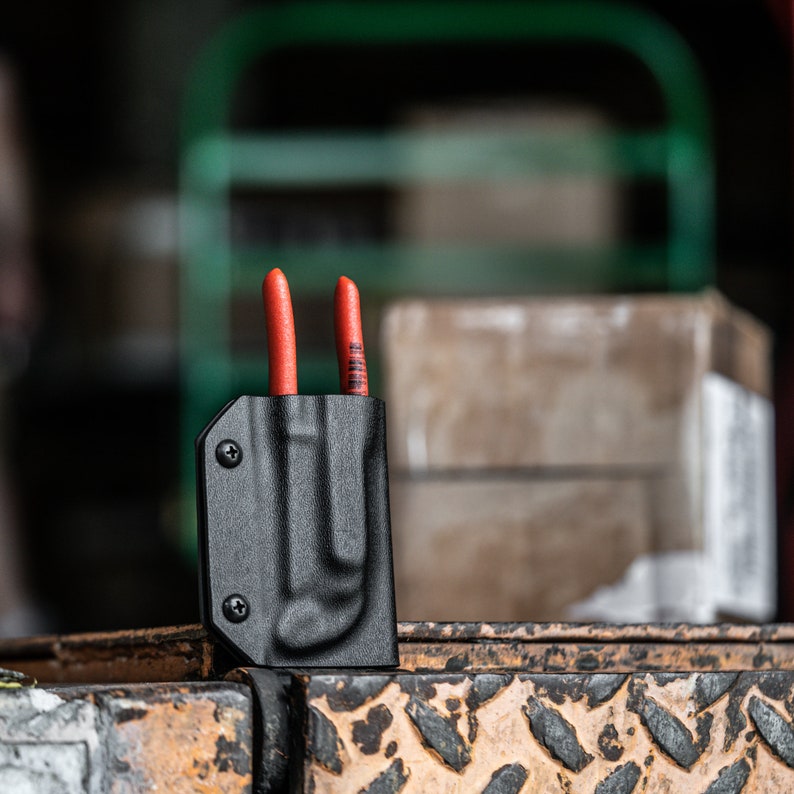 Puede incluir: Una funda de cuero negra con dos herramientas de mango rojo insertadas. La funda est&aacute; sobre una superficie texturizada.
