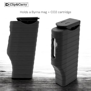 May include: Two black Clip&Carry Byrna mag holders. The holders are rectangular with a ridged design and a curved bottom. The top of the holder has a small, flat, rectangular lid. The text "Clip&Carry" is in the upper left corner. The text "Holds a Byrna mag + CO2 cartridge" is above the holders.