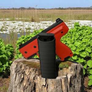 May include: A red and black paintball gun with a black magazine is displayed on a tree stump. The paintball gun has a textured grip and a black top. The magazine is cylindrical and black.