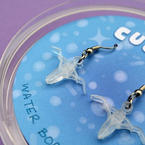 Op de afbeelding: Doorzichtige, transparante oorbellen met een uniek, abstract ontwerp. De oorbellen hangen aan zilverkleurige oorhaken. De oorbellen worden weergegeven op een blauwe achtergrond met de woorden "cute sea" en "water bo" zichtbaar.