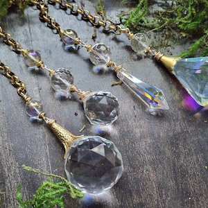 May include: Four decorative crystal pendants with gold-colored chains and accents. The pendants feature faceted crystal shapes, including spheres and a diamond-like prism. The crystals reflect light, creating a rainbow effect. The background includes green moss.