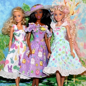 Peut inclure: Trois poupées vêtues de robes sur le thème de Pâques. Les robes sont blanches, lavande et bleu clair, ornées de motifs de lapins et d'œufs. Chaque poupée a des couleurs de cheveux et de peau différentes. Elles se tiennent devant une toile de fond peinte avec une scène de jardin.