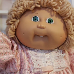 May include: A vintage doll with a round face, green eyes, and a small, open mouth revealing two teeth. The doll has light brown yarn hair and wears a pink floral dress with lace trim. The doll's arms are visible.