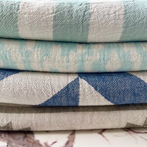 May include: A stack of four folded textiles with various patterns. The top textile has wide stripes of light green and white. The second has light blue and white stripes. The third has blue and white geometric shapes. The bottom textile has a gray star pattern.