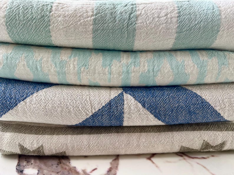 May include: A stack of four folded textiles with various patterns. The top textile has wide stripes of light green and white. The second has light blue and white stripes. The third has blue and white geometric shapes. The bottom textile has a gray star pattern.