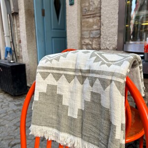 May include: A folded gray and white patterned blanket with fringe is draped over a bright orange chair. The blanket features a geometric design. The chair is outdoors, with a blue door and stone wall in the background.