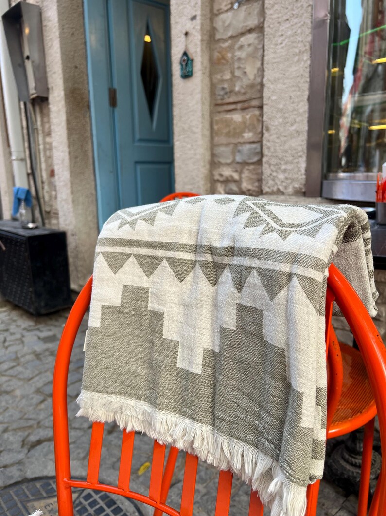 May include: A folded gray and white patterned blanket with fringe is draped over a bright orange chair. The blanket features a geometric design. The chair is outdoors, with a blue door and stone wall in the background.
