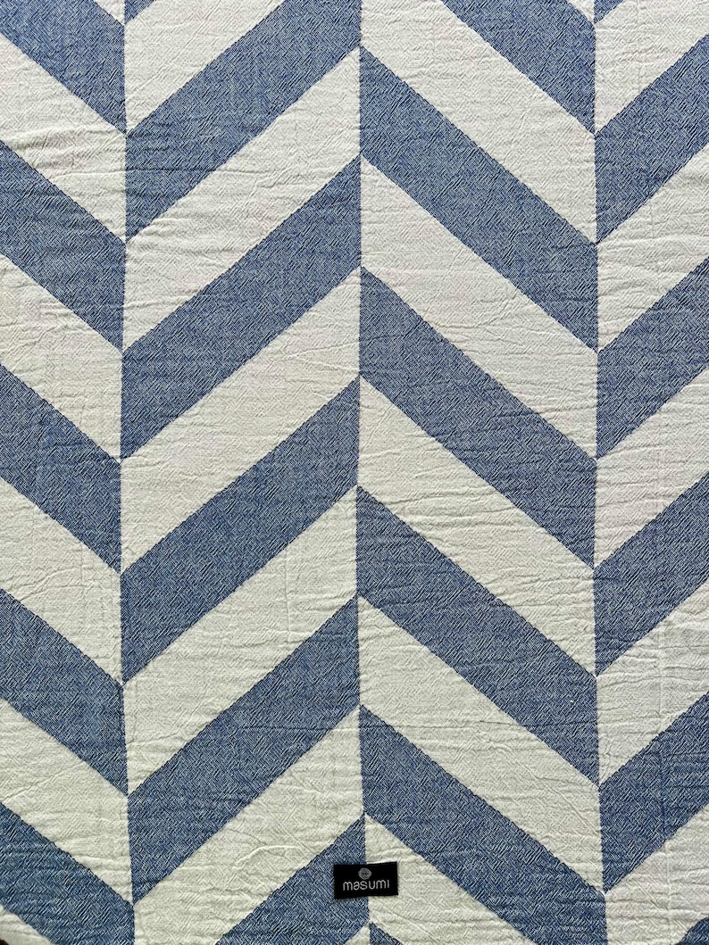May include: A blue and white chevron patterned textile. The design features alternating blue and white diagonal stripes, creating a zig-zag effect. The fabric has a slightly textured appearance. The brand name "masumi" is visible in the bottom right corner.