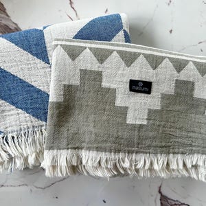 May include: Two folded cotton blankets. One has blue and white diagonal stripes, the other has a gray and white geometric pattern. Both have white fringe and a black label that reads "masumi".