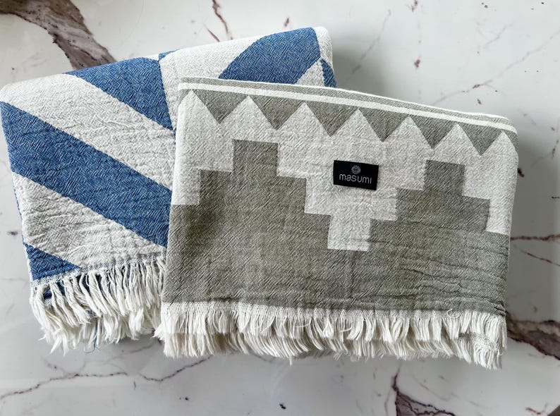 May include: Two folded cotton blankets. One has blue and white diagonal stripes, the other has a gray and white geometric pattern. Both have white fringe and a black label that reads "masumi".