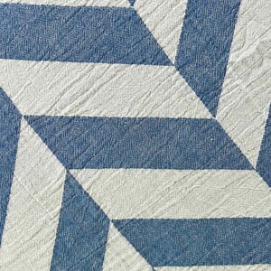 May include: Close-up of a textured textile with a geometric pattern. The design features alternating blue and white shapes, creating a modern, abstract look. The fabric has a woven appearance, with a tag in the bottom right corner.