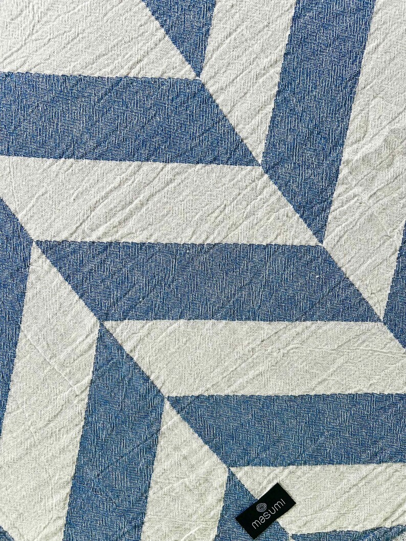 May include: Close-up of a textured textile with a geometric pattern. The design features alternating blue and white shapes, creating a modern, abstract look. The fabric has a woven appearance, with a tag in the bottom right corner.