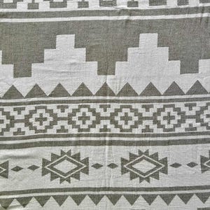 May include: A woven textile with a geometric pattern in white and gray. The design features repeating shapes, including diamonds, triangles, and stepped lines. The textile has a soft, textured appearance, suitable for a blanket or throw.