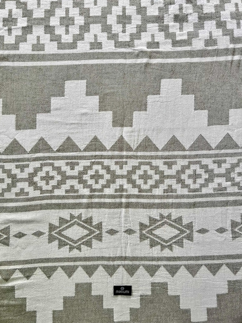 May include: A woven textile with a geometric pattern in white and gray. The design features repeating shapes, including diamonds, triangles, and stepped lines. The textile has a soft, textured appearance, suitable for a blanket or throw.