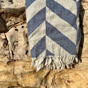 May include: A blue and white chevron patterned textile with a fringed edge. The fabric is draped over a rough, textured stone surface, creating a contrast between the soft textile and the rugged background.