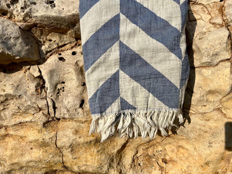 May include: A blue and white chevron patterned textile with a fringed edge. The fabric is draped over a rough, textured stone surface, creating a contrast between the soft textile and the rugged background.