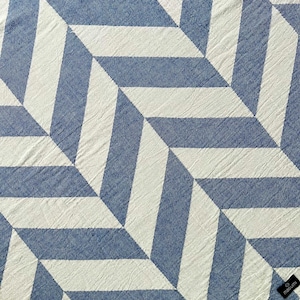 May include: A blue and white geometric patterned textile. The design features a repeating pattern of angled stripes, creating a three-dimensional effect. The textile is made of a textured fabric, with a small black tag in the corner.