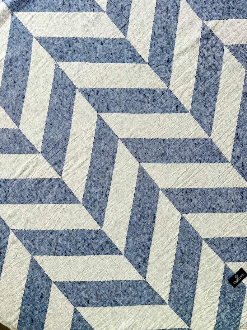 May include: A blue and white geometric patterned textile. The design features a repeating pattern of angled stripes, creating a three-dimensional effect. The textile is made of a textured fabric, with a small black tag in the corner.