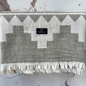 May include: A folded gray and white patterned towel with a geometric design and fringe. The towel has a black label that reads "masumi". The towel is made of a soft material.