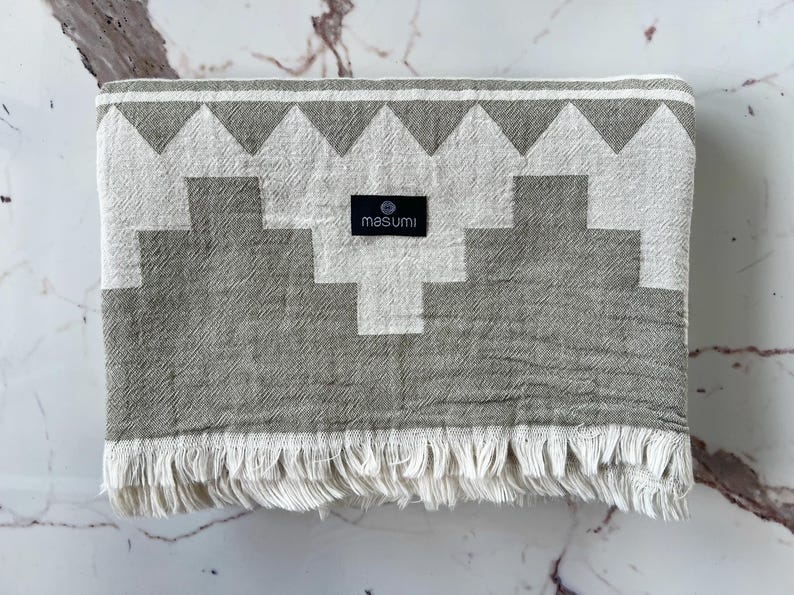 May include: A folded gray and white patterned towel with a geometric design and fringe. The towel has a black label that reads "masumi". The towel is made of a soft material.
