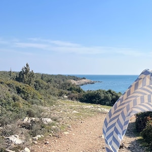May include: A scenic outdoor shot of a coastal path leading to the sea under a clear blue sky. A blue and white chevron patterned towel is blowing in the wind, adding a touch of color to the natural landscape.