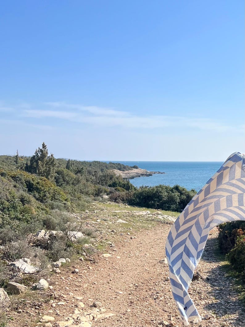 May include: A scenic outdoor shot of a coastal path leading to the sea under a clear blue sky. A blue and white chevron patterned towel is blowing in the wind, adding a touch of color to the natural landscape.