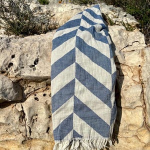 May include: A blue and white striped beach towel with a chevron pattern. The towel is made of a woven fabric and has a fringe along the bottom edge. The towel is draped over a light-colored rock formation.
