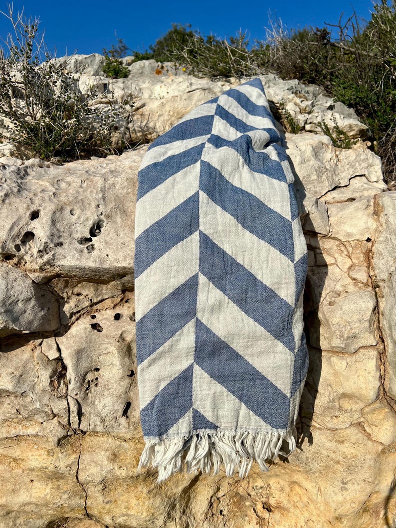May include: A blue and white striped beach towel with a chevron pattern. The towel is made of a woven fabric and has a fringe along the bottom edge. The towel is draped over a light-colored rock formation.