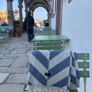 May include: A blue and white chevron patterned blanket with fringe is draped over a green chair. The blanket has a small black tag. The scene is outdoors, with a person walking in the background and green tables.