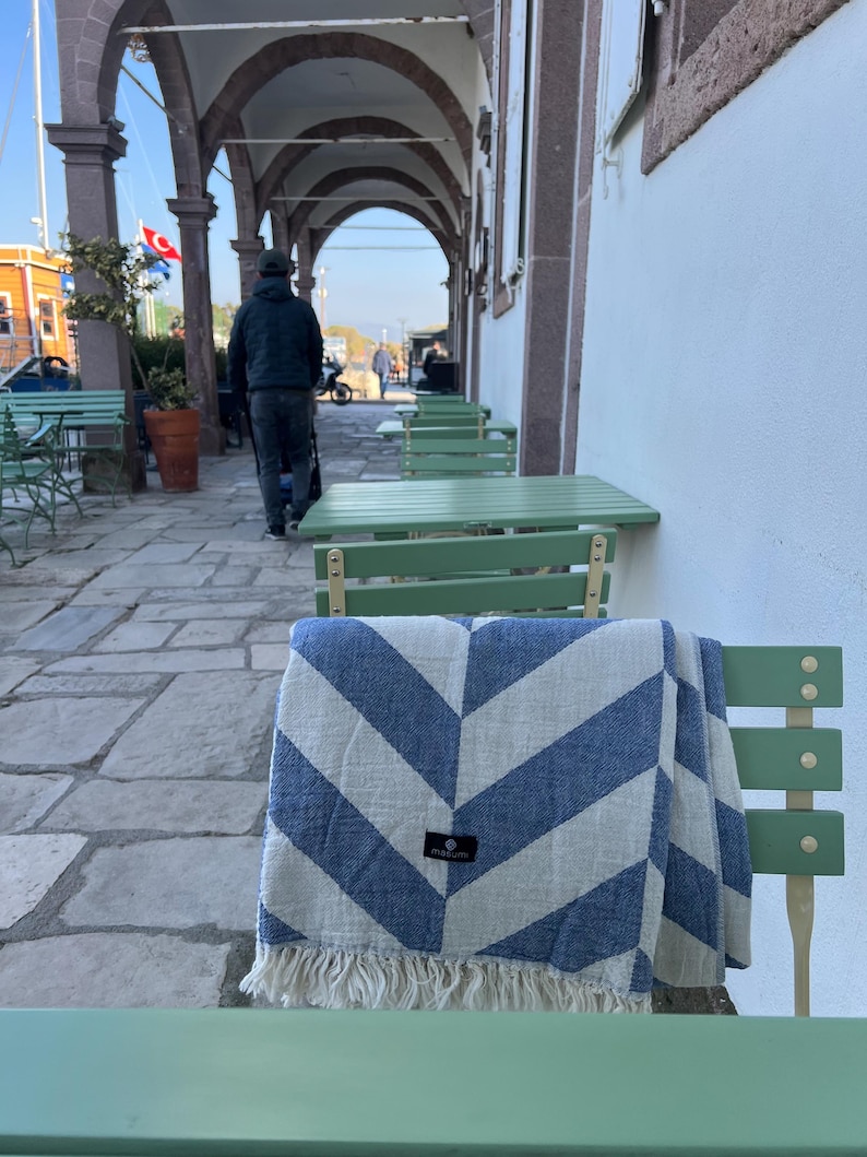 May include: A blue and white chevron patterned blanket with fringe is draped over a green chair. The blanket has a small black tag. The scene is outdoors, with a person walking in the background and green tables.