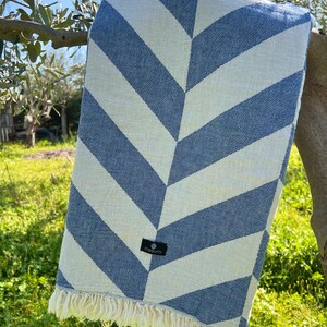 May include: A blue and white striped beach towel with a chevron pattern. The towel is hanging on a tree branch. The towel has a fringe on the bottom edge and a small black tag with the brand name.