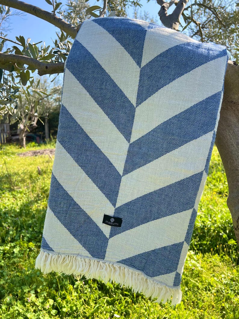 May include: A blue and white striped beach towel with a chevron pattern. The towel is hanging on a tree branch. The towel has a fringe on the bottom edge and a small black tag with the brand name.