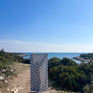 May include: A rectangular, blue and white patterned rug with a chevron design is displayed outdoors. The rug is hanging vertically against a backdrop of a blue sky, ocean, and coastal vegetation. The rug's dimensions are not visible.