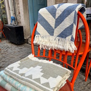 May include: A stack of folded blankets in various patterns and colors, including blue and white chevron, gray and white geometric, and light blue stripes. The blankets are displayed on an orange metal chair, showcasing their textures and designs.