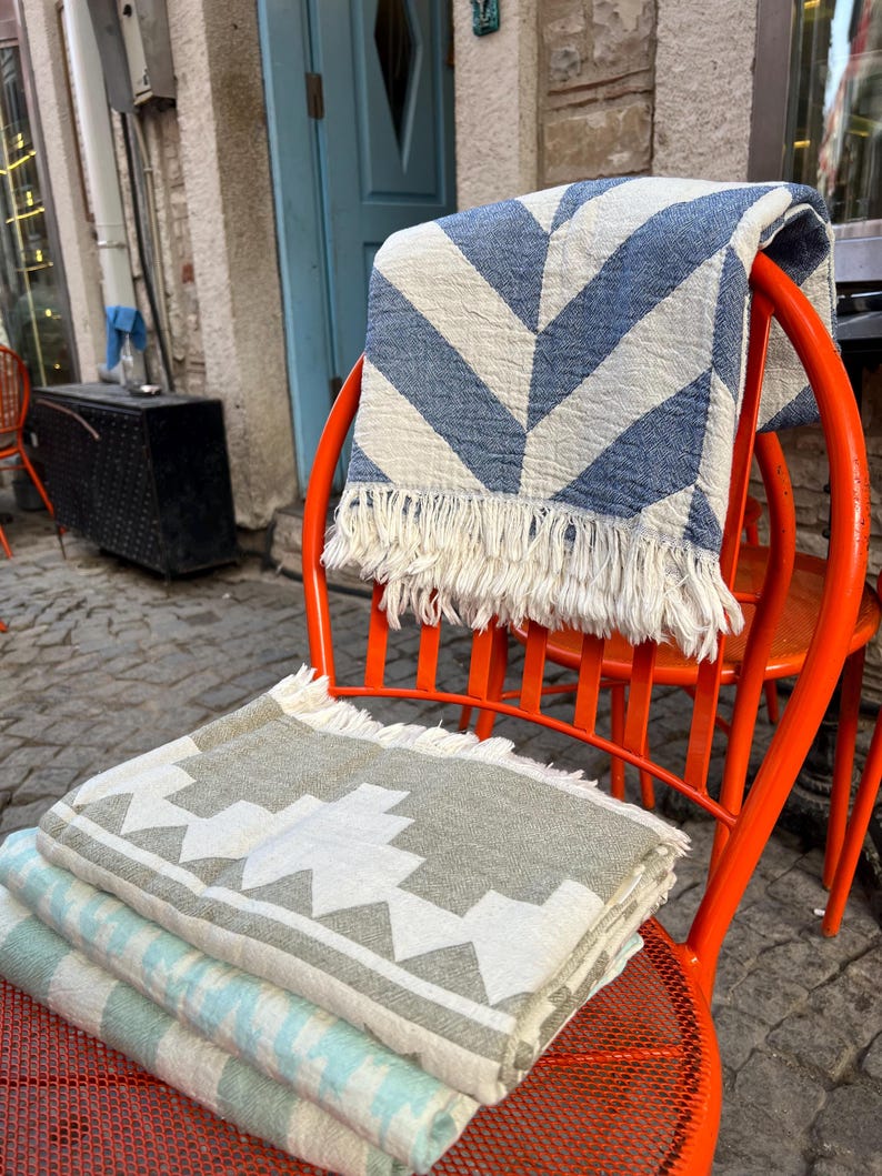 May include: A stack of folded blankets in various patterns and colors, including blue and white chevron, gray and white geometric, and light blue stripes. The blankets are displayed on an orange metal chair, showcasing their textures and designs.