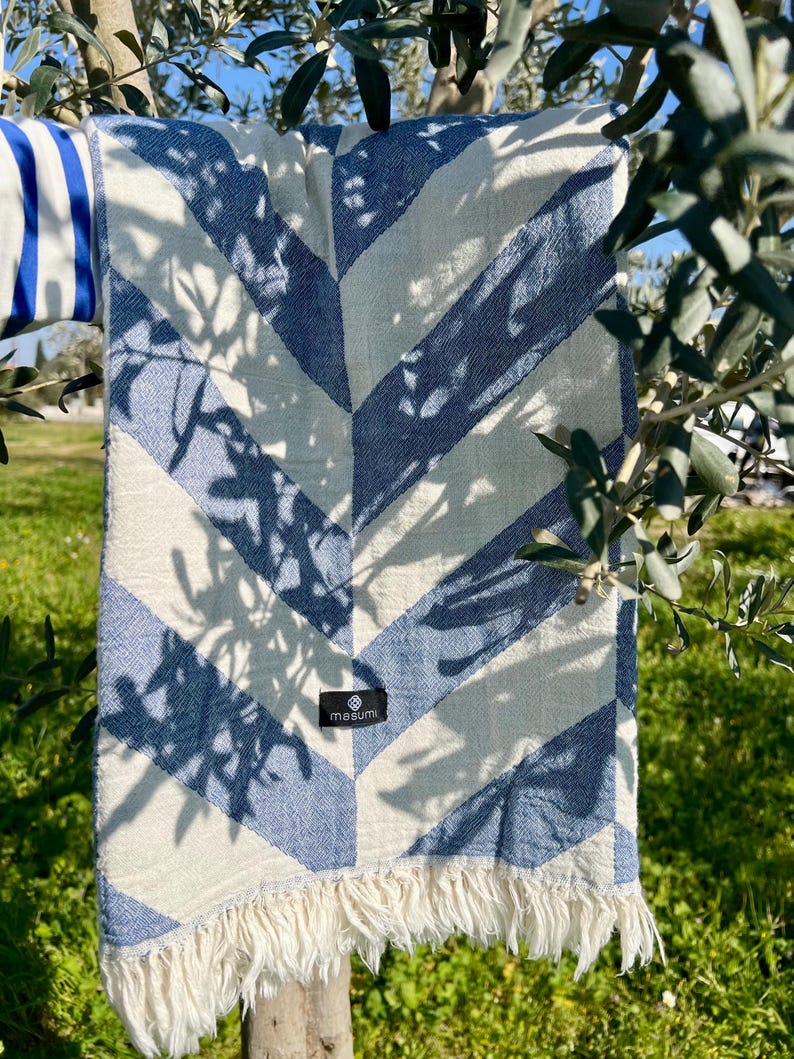 May include: A blue and white chevron patterned blanket with fringe. The blanket is draped over a tree branch. The blanket has a tag that says "masumi".
