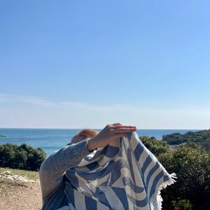 May include: A blue and white striped scarf with fringed edges. The scarf is draped over a person, with a clear blue sky and ocean in the background. The scarf's pattern is a series of diagonal stripes.