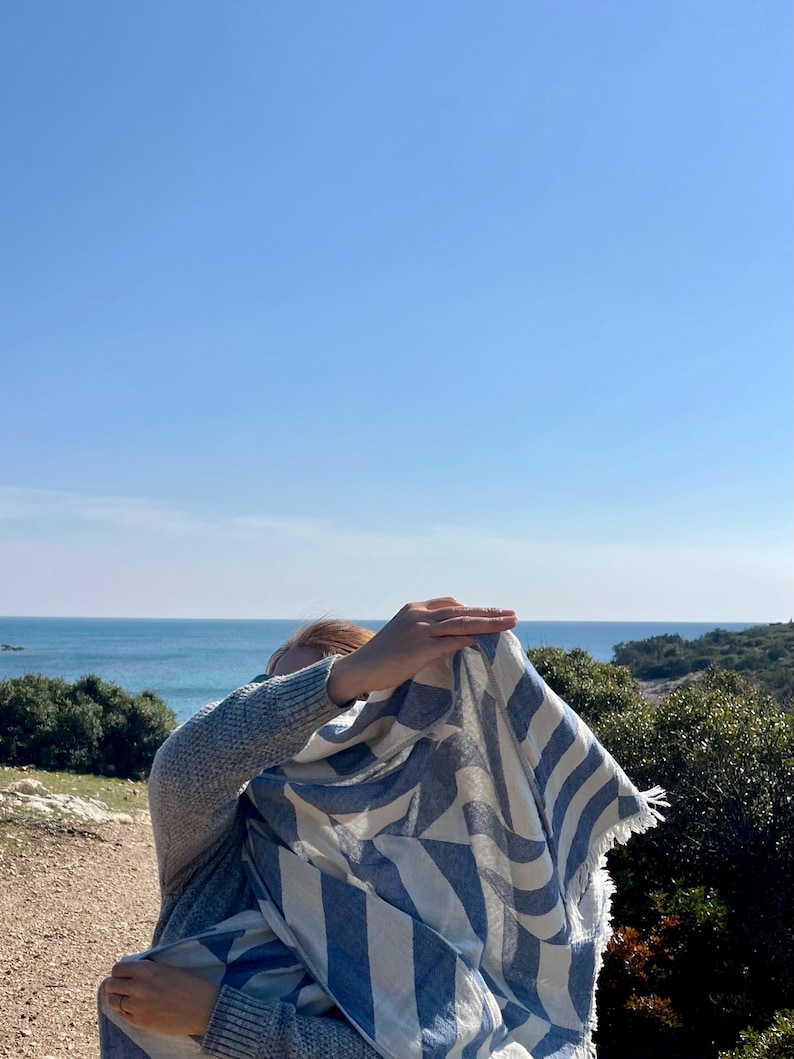 May include: A blue and white striped scarf with fringed edges. The scarf is draped over a person, with a clear blue sky and ocean in the background. The scarf's pattern is a series of diagonal stripes.