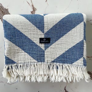 May include: A blue and white chevron patterned blanket with a fringed edge. The blanket has a tag that reads "masumi". The blanket is folded and appears to be made of a soft, woven material.