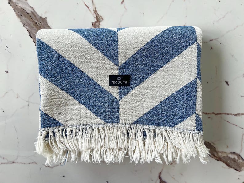 May include: A blue and white chevron patterned blanket with a fringed edge. The blanket has a tag that reads "masumi". The blanket is folded and appears to be made of a soft, woven material.