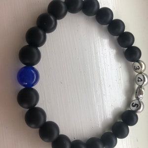 May include: Black beaded bracelet with a single blue bead and silver beads with the numbers 3, 4, and 5. The bracelet is made of matte black beads, creating a contrast with the blue and silver elements.
