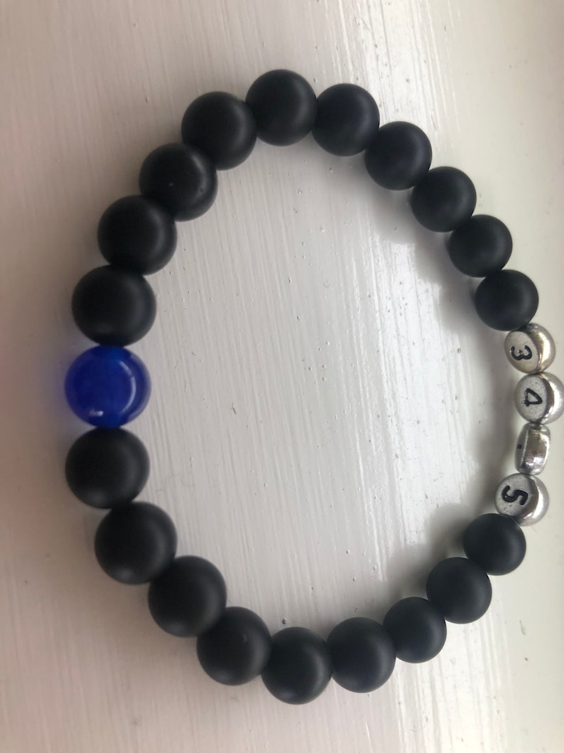 May include: Black beaded bracelet with a single blue bead and silver beads with the numbers 3, 4, and 5. The bracelet is made of matte black beads, creating a contrast with the blue and silver elements.