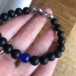 May include: Black beaded bracelet with silver letter beads spelling "SAS" and a single blue bead. The bracelet is made of matte black beads and is held in a hand.