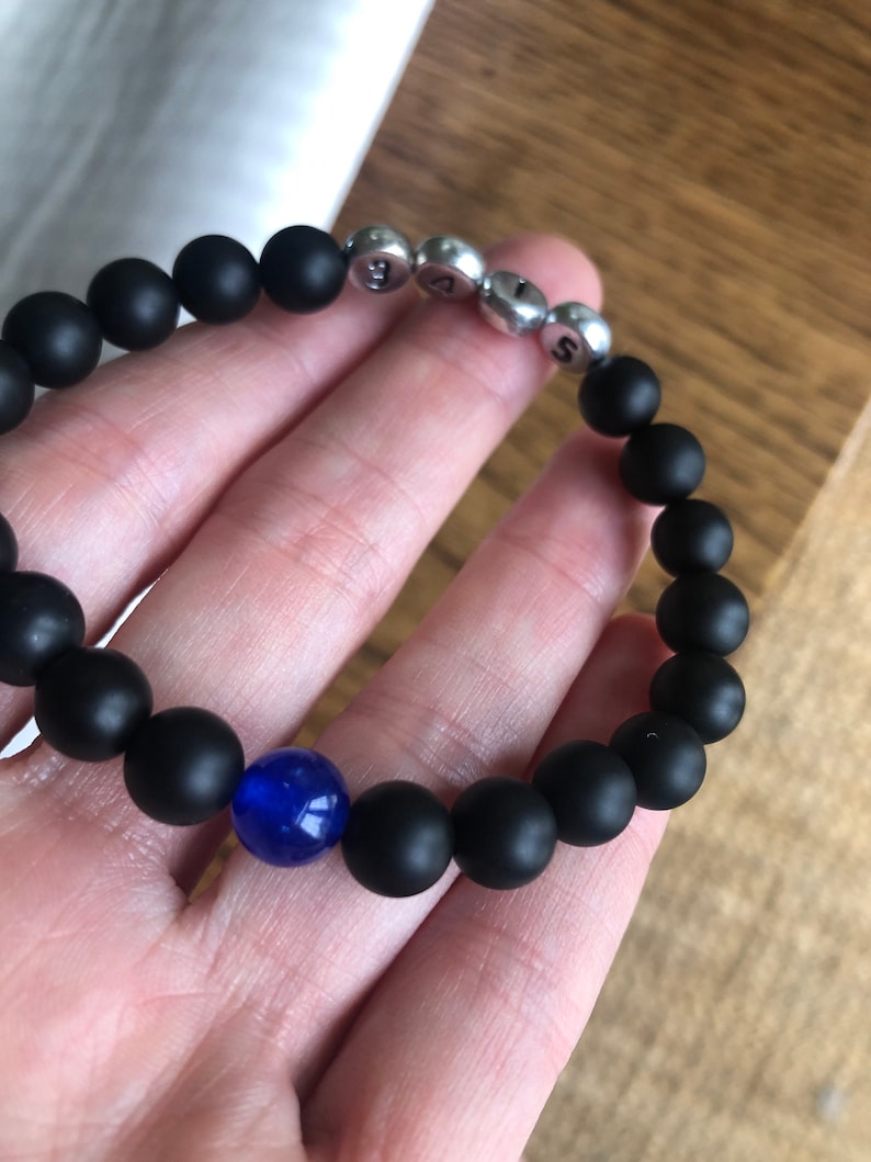 May include: Black beaded bracelet with silver letter beads spelling "SAS" and a single blue bead. The bracelet is made of matte black beads and is held in a hand.
